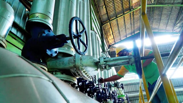 Industrial technician operating a large steel valve wheel in a factory with complex piping and safety gear for maintenance and plant operations