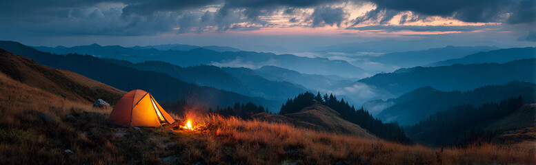 Fototapeta premium A scenic vista shows an illuminated tent on a hillside as a campfire burns. The background features layers of hazy mountains. The twilight sky