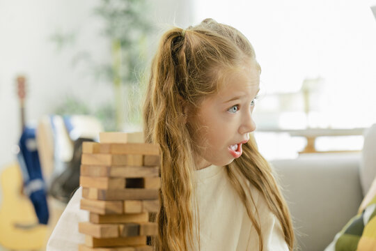 A young girl looks shocked while playing a game of wooden block stacking, reacting to a critical moment in the game.