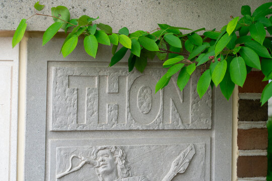 THON engraved stone relief featuring a musician playing a lute, adorned with vibrant green foliage and small red berries, set against a textured concrete wall, symbolizing enduring spirit and
