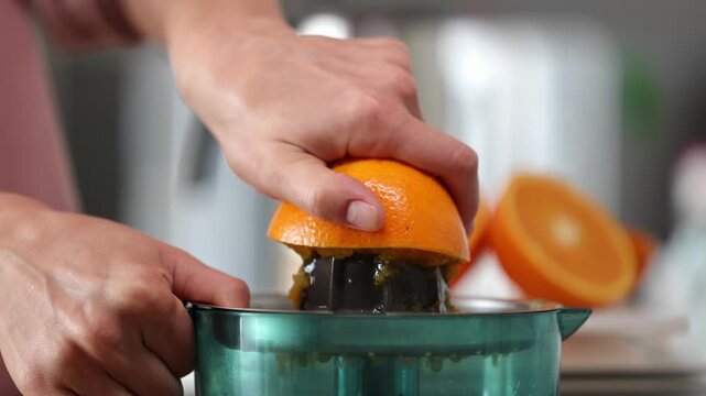 Female hand presses orange onto juicer, extracting juice while additional oranges are visible in the background on a kitchen countertop