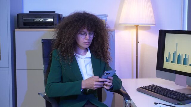 Businesswoman with curly hair and glasses using smartphone at office desk with computer showing bar chart, printer and lamp in background, modern workplace, blue lighting.