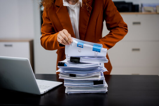 Businesswoman handling a thick stack of paperwork on a desk beside a laptop in an office, symbolizing bureaucracy, workload, and document management.