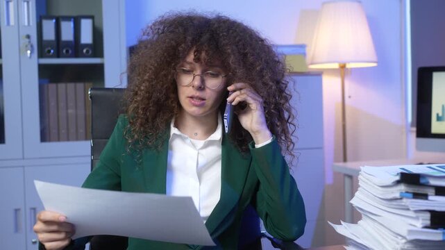 Curly-haired businesswoman in glasses working at desk, talking on phone while reviewing document in modern office with lamp, computer, shelves and stacks of paperwork.