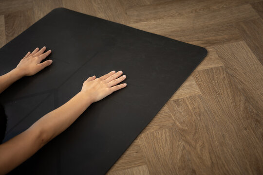 Calm person placing hand and arm on black yoga mat over wood floor during fitness and health routine feeling peaceful and focused while resting in studio