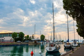 Sailboats are moored in the marina at Peschiera del Garda, with reflections on the water and trees lining the shore under a cloudy sky © Nizoli