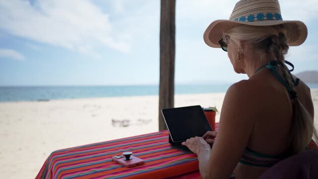 Happy mature woman using computer tablet, and phone at a beach hut during her tropical vacation by the sea.