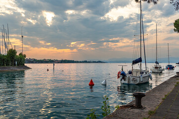 Scenic view of sailboats moored in calm waters at sunset, with a shoreline featuring trees and distant mountains in Peschiera del Garda, Italy © Nizoli