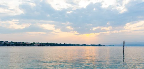 Scenic view of Lago Garda in Italy featuring calm waters, distant shoreline, and a cloudy sky during sunset, reflecting soft colors on the water surface © Nizoli