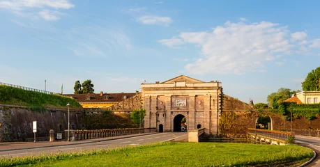 Porta Verona, a historic fortress entrance in Peschiera del Garda, surrounded by greenery and a winding road under a cloudy sky at sunrise © Nizoli
