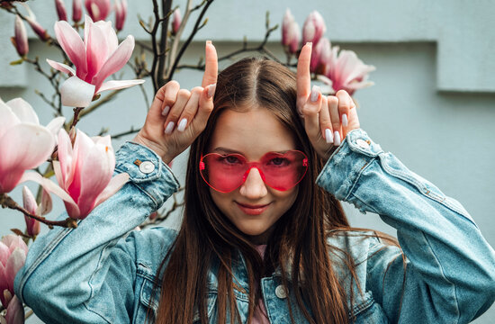 Smiling girl in pink heart glasses making horn gesture by a magnolia