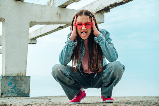 Playful rebellious girl in denim and sunglasses crouching by the sea
