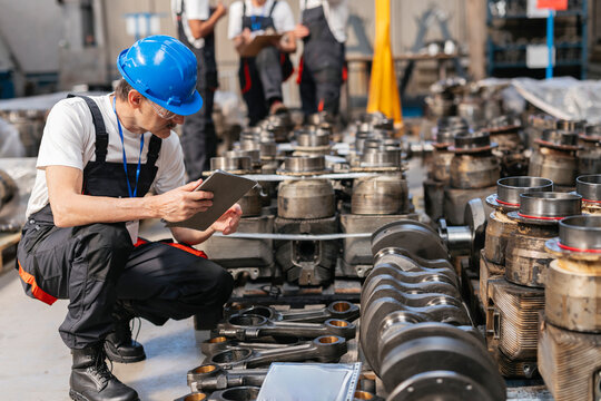Engineer kneeling in factory inspecting engine part with tablet