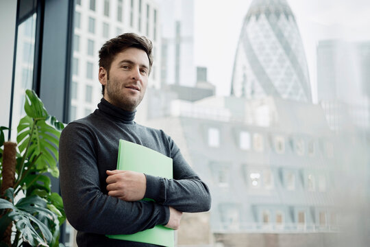 Mature businessman holding file folder near glass wall at office