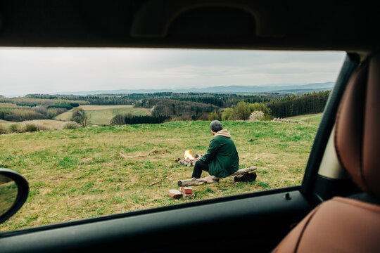 Camper sitting by a fire in the countryside viewed from a car