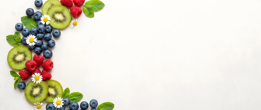 Top view of fresh kiwi slices, raspberries and blueberries with mint leaves and chamomile flowers forming a crescent frame on light textured background