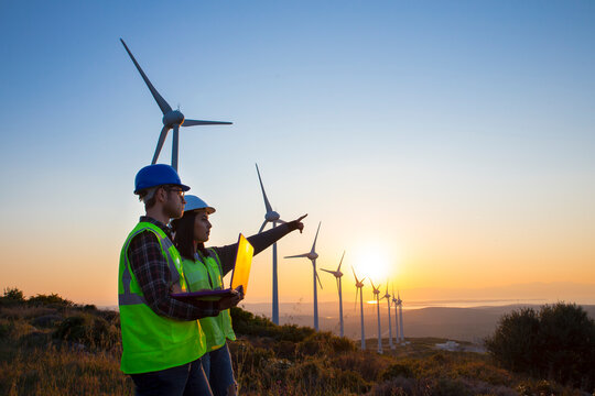The engineers are reviewing data on a tablet, analyzing the performance of wind turbines in a field. indicating their role in maintaining the renewable energy infrastructure.