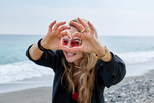 Blonde woman smiling making heart shape with hands by the sea