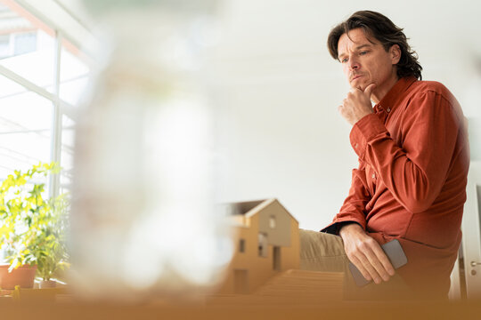 Focused architect sitting with hand on chin and looking at model house in home office