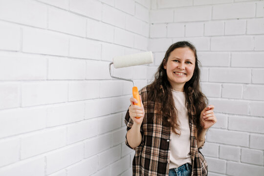 Smiling woman holding a paint roller by a white brick wall indoors
