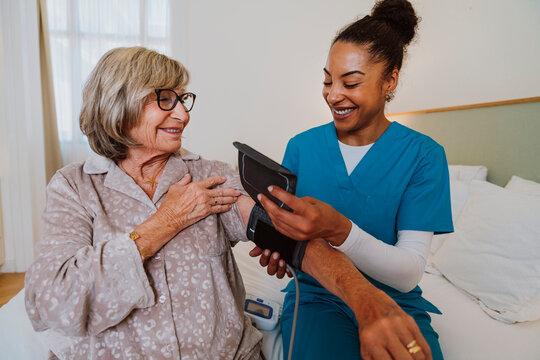 Nurse checking blood pressure of senior woman at home in pajamas