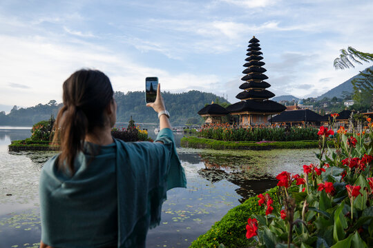Traveler taking photo of Bali temple by lake with flowers and mountains