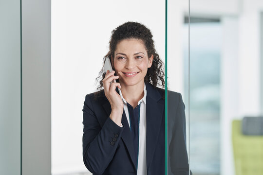 Happy businesswoman talking on smart phone near glass door in office