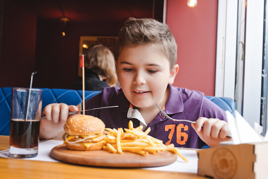 Young boy using a knife and fork to eat a burger and french fries on a wooden board in a restaurant.