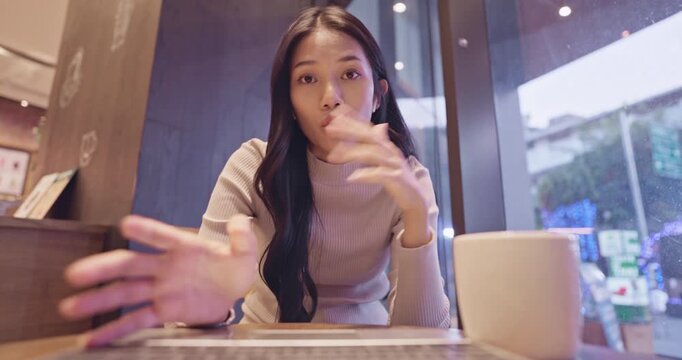 Young Asian woman with long hair in a cozy knit sweater talks excitedly with hand gestures at a stylish cafe table.