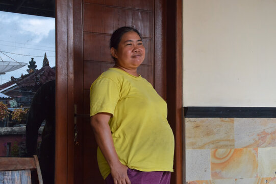 A bright smiling Asian woman standing comfortably in front of a wooden door panel at the house veranda during a calm day.