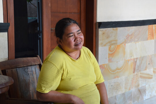 A pleasant middle aged Asian woman wears a yellow shirt while sitting relaxed and smiling gently on a wooden chair on her house veranda.