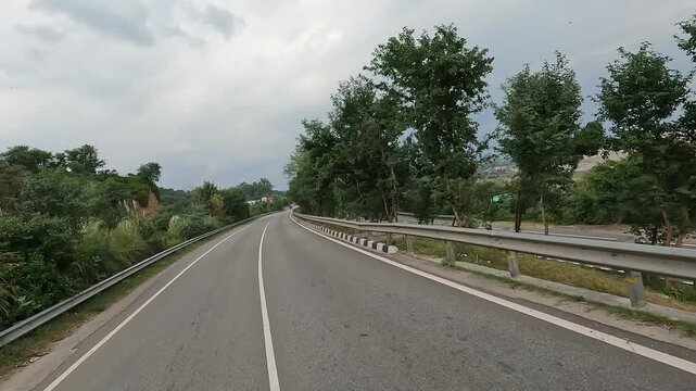 4K footage of a scenic curved road and bridge passing through a green hilly landscape under a cloudy sky, with open riverbed and countryside views.