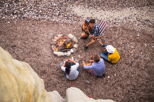 Group of friends sitting on beach with guitar playera campfire in a  outdoor setting, sitting on beach with guitar player.
