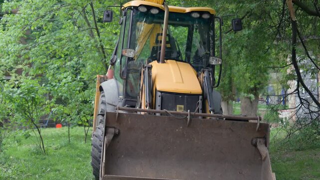Yellow Backhoe Loader Parked Among Green Trees in Urban Yard | Front Bucket Close-Up View | Heavy Construction Equipment Idle Outdoors