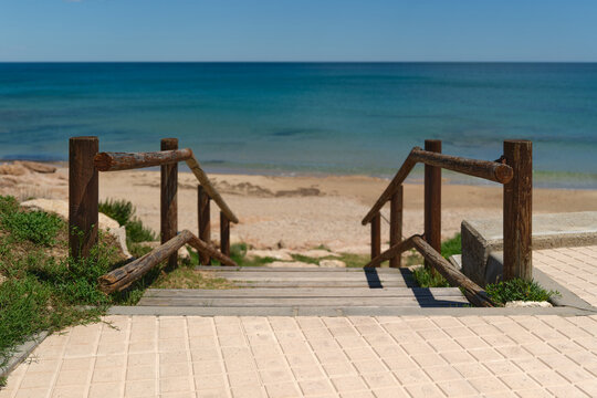 Wooden beach access stairs leading through dunes to a sandy Mediterranean shoreline with calm blue sea. Sunny coastal landscape with natural summer atmosphere and seaside vacation mood.