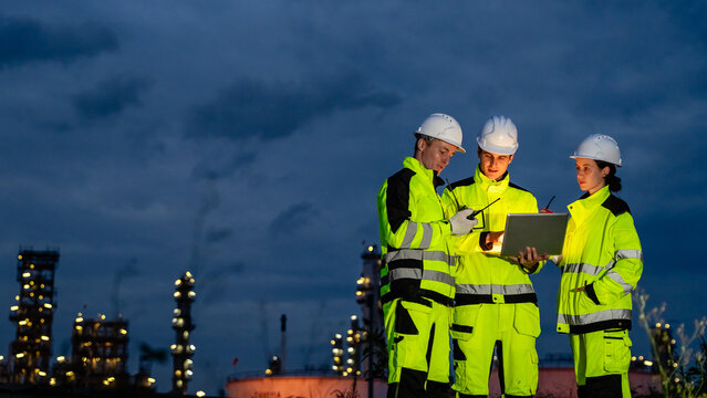 Professional industrial engineer team collaborating with laptop at night shift illuminated petrochemical refinery plant for sustainable corporate oil and gas manufacturing business strategy.