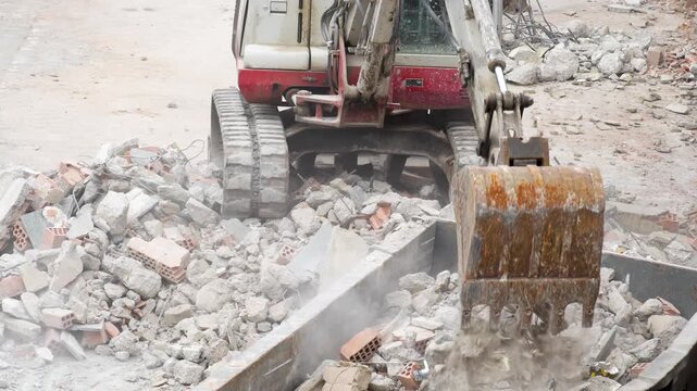 Tracked excavator loads dusty rubble, broken bricks and concrete into a metal skip during demolition cleanup at an active construction site.