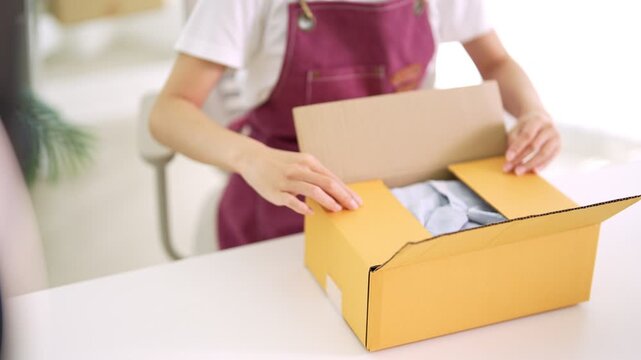 Asian woman live streaming online merchant packing yellow cardboard box with clothing preparing order shipment for ecommerce business home delivery service