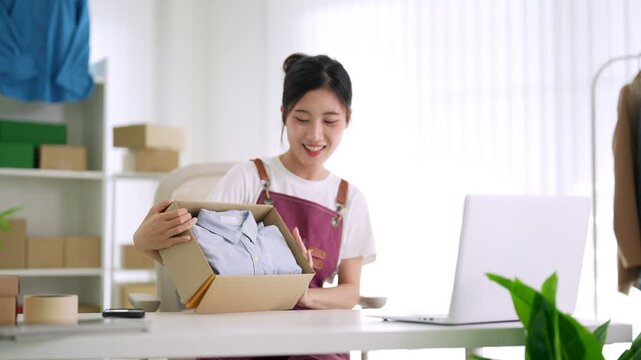 Asian women small business owner packing product box live streaming online selling clothing merchandise presenting garment shirt home office workspace