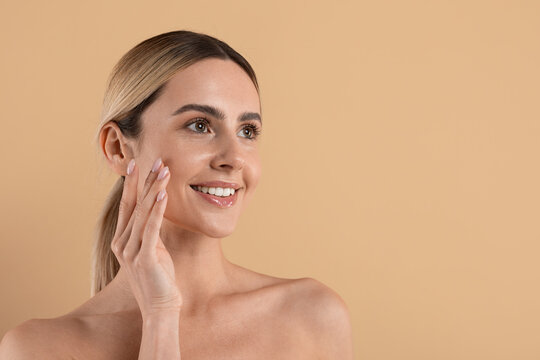 Smiling young caucasian woman with radiant healthy skin touching cheek in beauty and skincare concept isolated on clean beige studio background, copy space