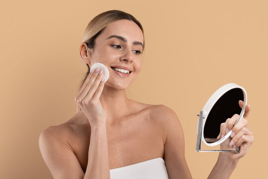 Beautiful smiling woman removing makeup with cotton pad while looking in handheld mirror during skincare routine, standing on beige studio background