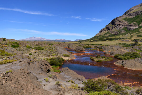 Paisaje del r&iacute;o Agrio con lecho rojizo, monta&ntilde;as, vegetaci&oacute;n de estepa y el volc&aacute;n Copahue al fondo bajo un cielo azul en la Patagonia.