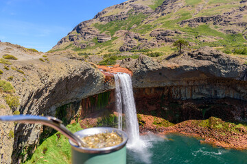 Primer plano de un mate tradicional con bombilla enfocado, con la cascada Salto del Agrio y montañas desenfocadas al fondo bajo un cielo despejado. Paisaje turístico de la Patagonia Argentina © SeVPH