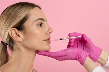 Side profile portrait of beautiful young woman during cosmetic lip enhancement procedure with syringe and beautician hands on pink studio background © Anastasiya
