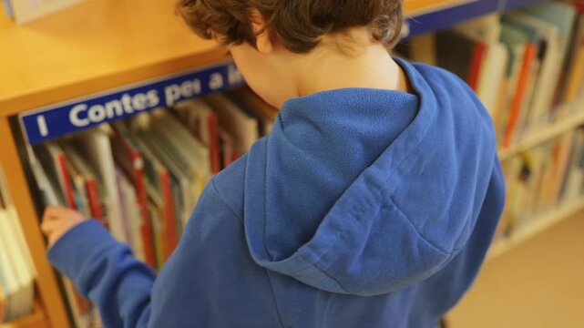 Young student browsing books on a bookshelf in a public school library, focused on selecting a children's storybook for reading and enhancing his childhood education