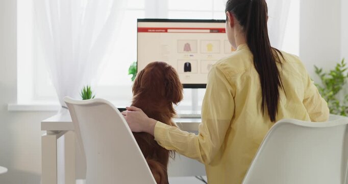 Dog and owner browsing online shopping on computer. At home a woman sits with her pet as they view the screen together. Clean workspace enables simple checkout. Pet and owner at home.