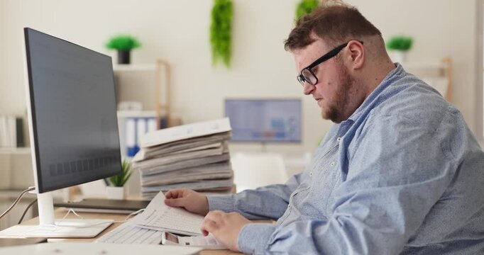 Overweight man accountant in office with paperwork. Working at a computer, he reviews bills and prepares budget analysis for small finance tasks. Concept shows stress and tired work overload.