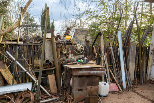 Backyard storage area filled with scrap metal, pipes, wooden boards, tools, rusty parts, plastic containers, fencing, branches, and construction leftovers in an outdoor workshop setting.