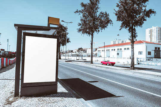 Blank vertical advertising panel at modern bus shelter on sunny urban street, editable outdoor display template with blue sky, city buildings and red convertible car