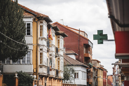 Historic yellow townhouse with white wooden glass balconies, tiled roof, flags and pharmacy cross, charming street architecture in Asturias, Spain